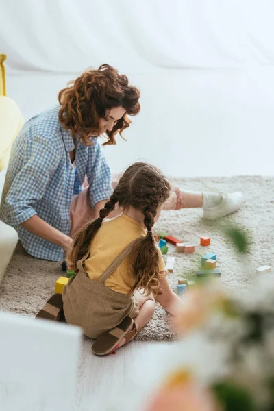 Selective focus of babysitter and kid sitting on floor and playing with multicolored blocks — Stock Photo