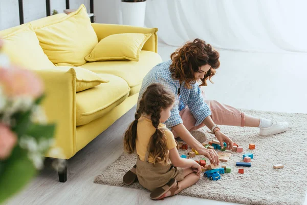 Foyer sélectif de jeune nounou et enfant assis sur le sol et jouer avec des blocs multicolores — Photo de stock