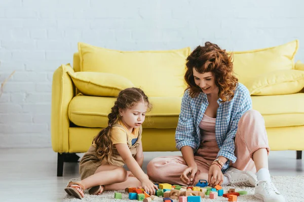Улыбающаяся няня и милый ребенок, сидящий на полу и играющий с разноцветными блоками — стоковое фото
