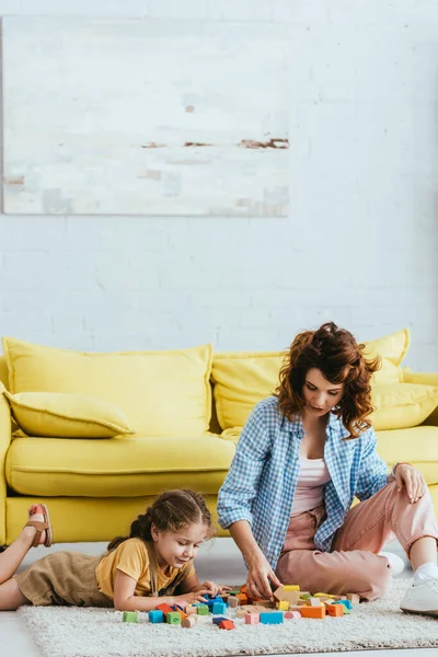 Junges Kindermädchen und entzückendes Kind, das mit bunten Klötzen auf dem Boden neben dem gelben Sofa spielt — Stockfoto