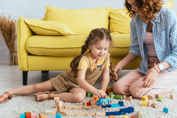 Young nanny sitting near adorable kid playing with toy car near multicolored blocks on floor — Stock Photo