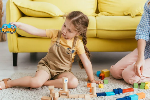 Cropped view of babysitter near child holding toy car while sitting on floor near multicolored blocks — Stock Photo