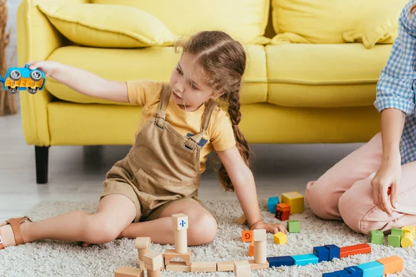 Vista cortada de babá perto de criança segurando carro de brinquedo enquanto sentado no chão perto de blocos multicoloridos — Fotografia de Stock