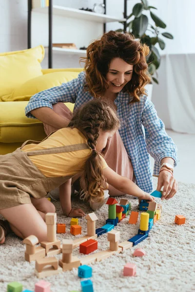 Улыбающаяся няня с ребенком играет с разноцветными блоками, сидя на полу — стоковое фото