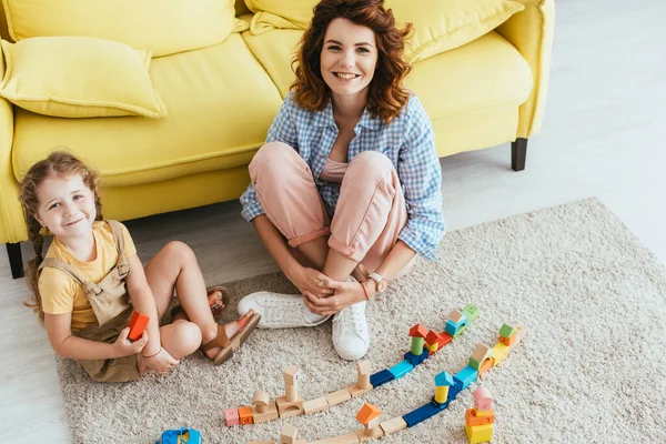 Vista ad alto angolo di infermiera felice e bambino guardando la fotocamera mentre seduto sul pavimento vicino scatola di cartone e blocchi multicolore — Foto stock