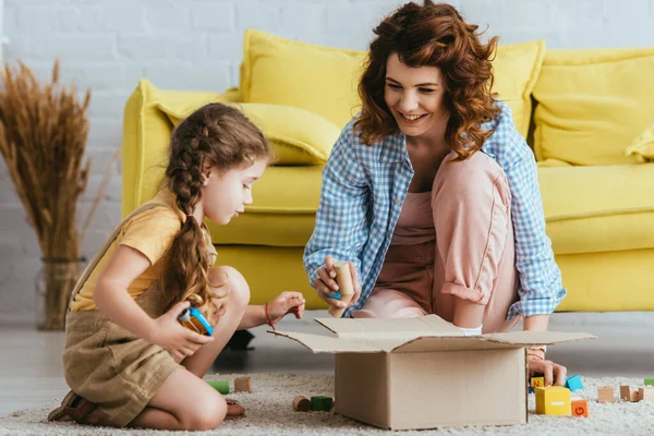Babysitter sorridente e bambino carino seduto sul pavimento vicino alla scatola di cartone con blocchi multicolori — Foto stock
