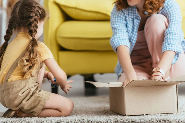 Cropped view of babysitter and kid sitting on floor near cardboard box — Stock Photo