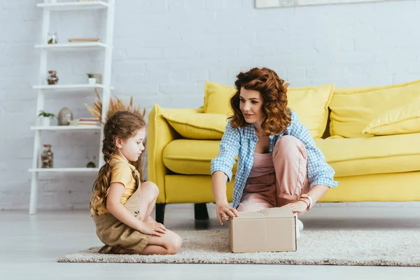Young nanny and adorable kid sitting on floor near cardboard box — Stock Photo