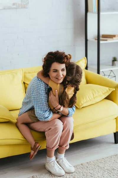 Glücklich Kindermädchen umarmt Kind, während es auf gelbem Sofa sitzt — Stockfoto