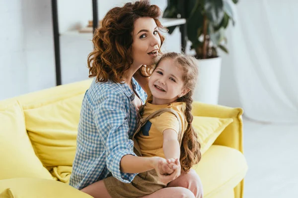 Feliz niñera y adorable niño cogido de la mano y sonriendo a la cámara mientras está sentado en el sofá - foto de stock