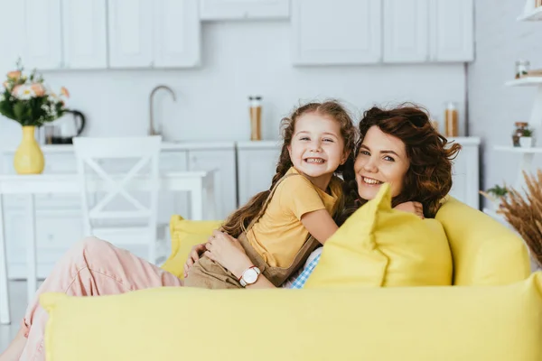 Niñera alegre y el niño sonriendo a la cámara mientras se abraza en el sofá en la cocina - foto de stock