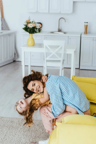 Souriant nounou tenant heureux enfant à l'envers tout en s'amusant sur canapé dans la cuisine — Photo de stock