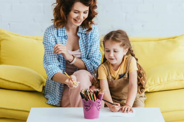 Happy nanny and child drawing with pencils while sitting on yellow sofa — Stock Photo