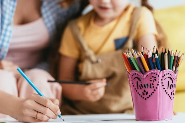 Selective focus of nanny drawing with pencil near child and pen holder on table — Stock Photo