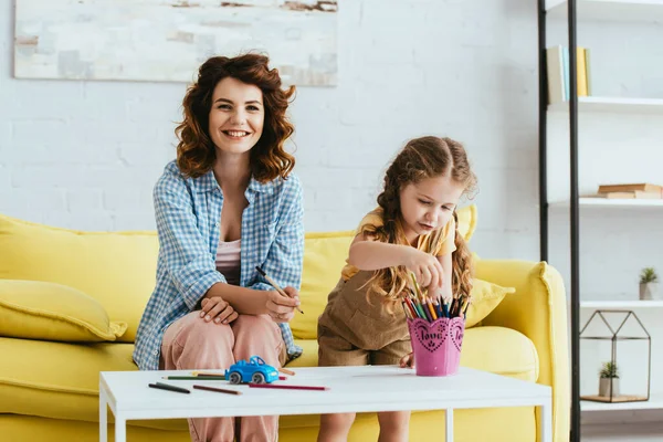 Tata felice sorridente alla macchina fotografica mentre adorabile bambino che prende la matita dal portapenne — Foto stock