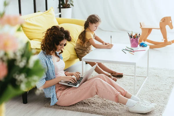 Messa a fuoco selettiva di babysitter seduto sul pavimento e lavorare sul computer portatile vicino disegno bambino con matita — Foto stock