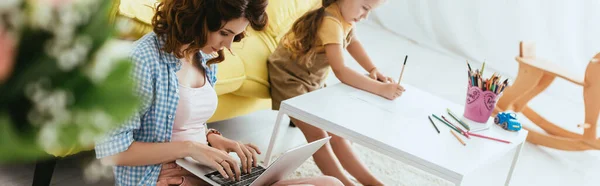 Enfoque selectivo de la niñera joven que trabaja en el ordenador portátil cerca del dibujo del niño con lápiz, imagen horizontal - foto de stock