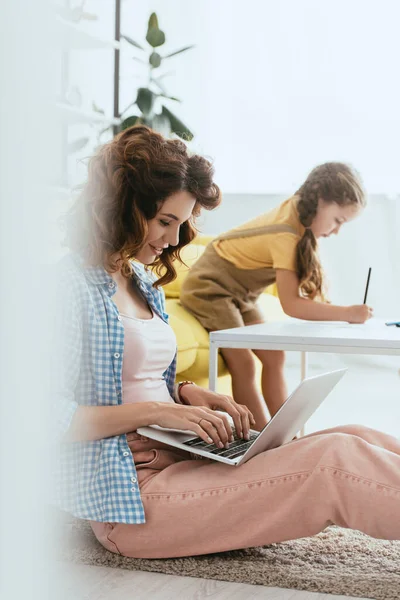 Selective focus of smiling babysitter working on laptop near kid drawing with pencil — Stock Photo