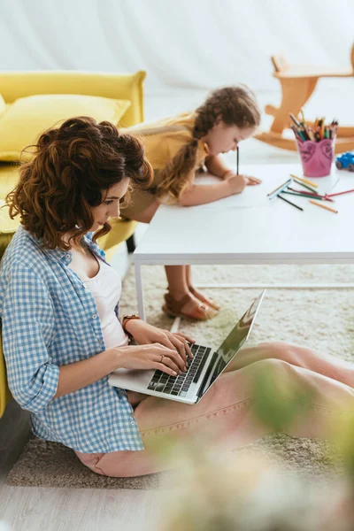 Foyer sélectif de baby-sitter travaillant sur ordinateur portable près de dessin d'enfant avec des crayons — Photo de stock