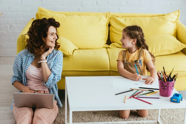 Happy child drawing with pencils while looking at nanny working on laptop — Stock Photo
