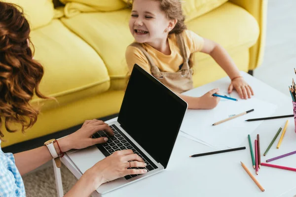 Vue grand angle du dessin heureux de l'enfant avec crayon près de nounou en utilisant un ordinateur portable avec écran vide — Photo de stock