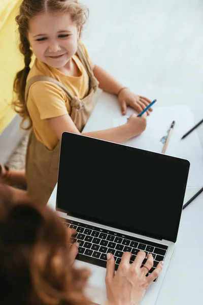 Вид сверху на няню с помощью ноутбука с чистым экраном рядом с счастливым ребенком рисунок с карандашом, избирательный фокус — стоковое фото