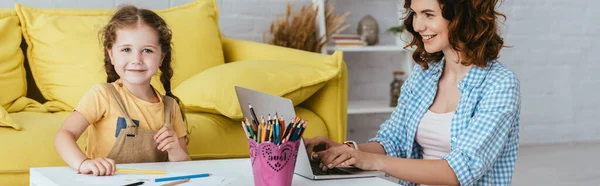 Panoramic shot of happy child drawing near smiling babysitter working on laptop — Stock Photo
