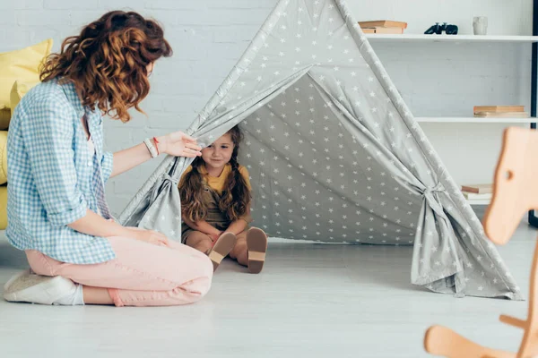 Young nanny looking at adorable child sitting in kids wigwam — Stock Photo