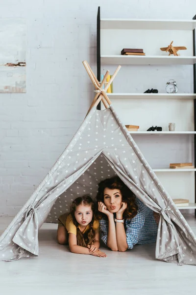 Lindo niño y niñera joven haciendo caras de pato y mirando a la cámara mientras está acostado en los niños wigwam - foto de stock