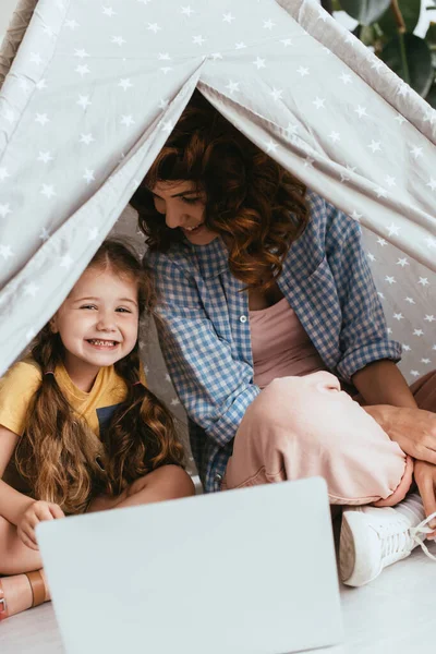 Niñera feliz y adorable niño sentado en juguete wigwam cerca de la computadora portátil - foto de stock