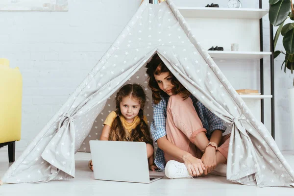 Young nanny sitting with crossed legs near cute kid using laptop in toy wigwam — Stock Photo