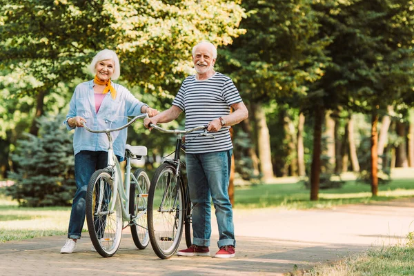 Casal de idosos sorrindo para a câmera enquanto caminhava com bicicletas na passarela no parque — Fotografia de Stock
