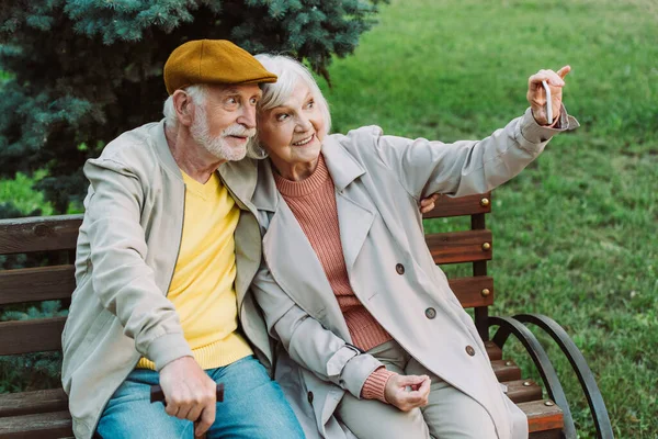 Pareja mayor sonriendo mientras toma selfie con teléfono inteligente en el banco en el parque - foto de stock