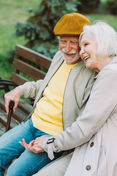 Sorridente coppia anziana che si tiene per mano mentre si siede sulla panchina nel parco — Foto stock