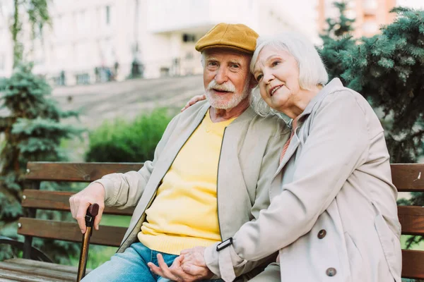 Selective focus of senior man looking at camera while sitting near smiling wife on bench in park — Stock Photo