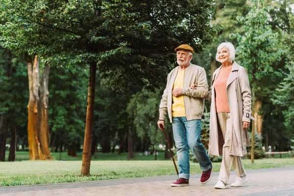 Улыбающаяся пожилая женщина, идущая рядом с мужем по дорожке в парке — стоковое фото