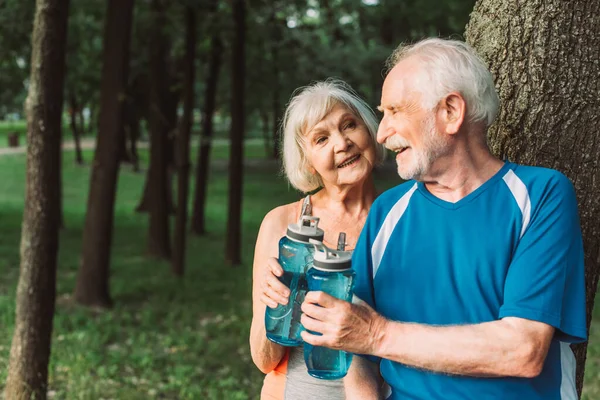 Smiling senior couple holding sports bottles in park — Stock Photo