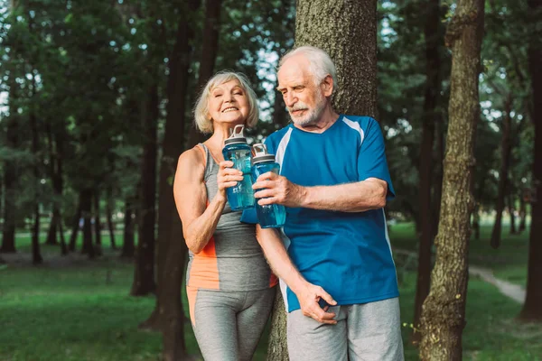 Sorridente donna anziana che tiene una bottiglia sportiva vicino al marito e all'albero nel parco — Foto stock