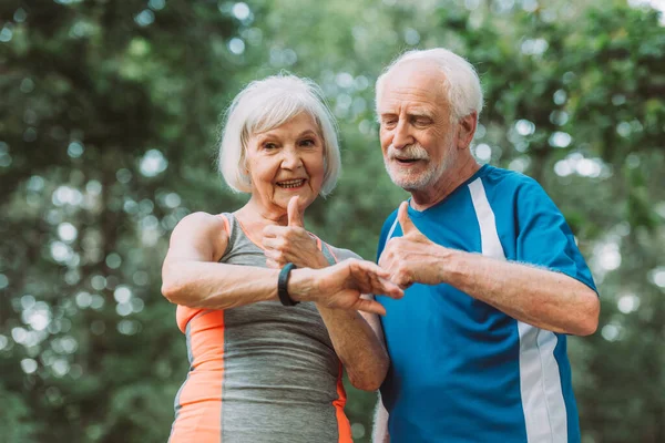 Sonriendo pareja mayor mirando smartwatch y mostrando pulgares hacia arriba en el parque - foto de stock