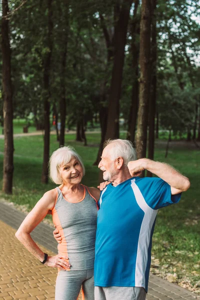 Улыбающаяся пожилая женщина обнимает мужа в спортивной одежде в парке — стоковое фото