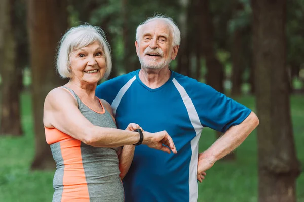 Mujer mayor sonriente con rastreador de fitness sonriendo a la cámara cerca del marido en el parque - foto de stock