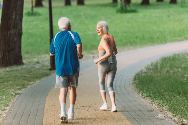 Вид ззаду усміхненої старшої жінки, яка дивиться на чоловіка під час бігу в парку — стокове фото