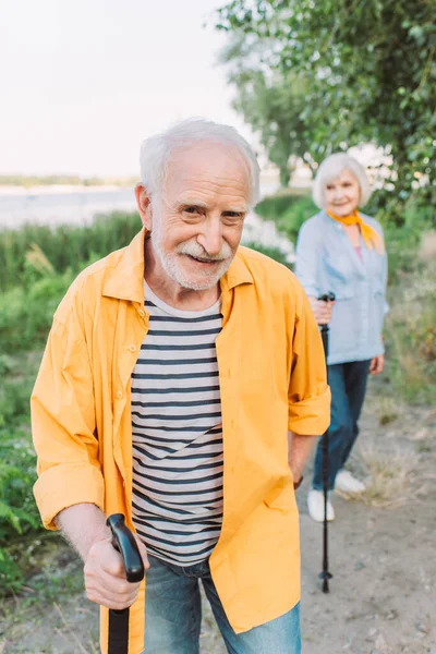 Вибірковий фокус усміхненого старшого чоловіка з ходьбою палицею, дивлячись на камеру біля дружини в парку — стокове фото