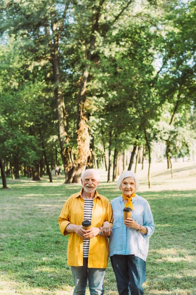 Alegre pareja de ancianos con vasos de papel mirando a la cámara en el parque - foto de stock