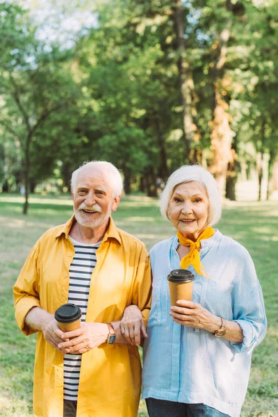 Mujer sonriente sosteniendo café para ir y sonriendo a la cámara cerca de marido mayor en el parque - foto de stock