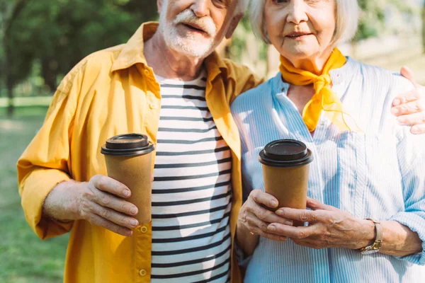 Vista cortada do homem sênior segurando café para ir e abraçando a esposa no parque — Fotografia de Stock