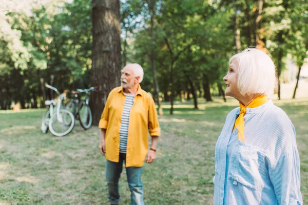Foco seletivo de sorrir mulher idosa olhando para longe perto do marido no parque — Fotografia de Stock