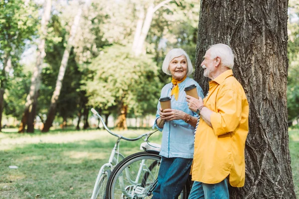 Enfoque selectivo del hombre mayor sonriente sosteniendo una taza de papel cerca de la esposa y las bicicletas en el parque - foto de stock