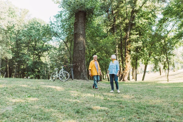 Улыбающаяся пожилая пара, гуляющая по газону с велосипедами возле дерева в парке — стоковое фото