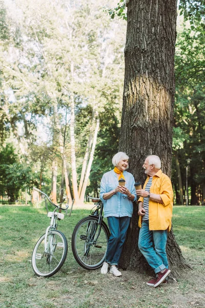 Elderly couple smiling at each other while holding paper cups near tree and bikes in park — Stock Photo